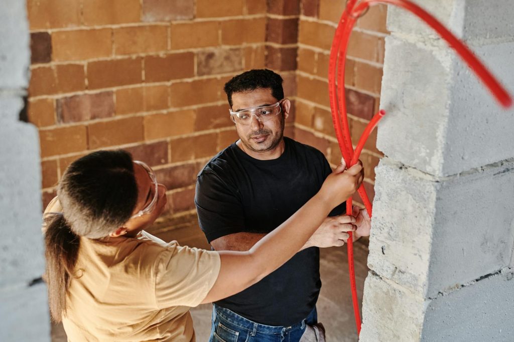 Two construction workers fitting red tubing on a brick wall at a construction site